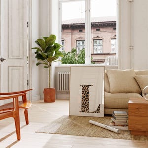 May include: A framed art print featuring the letter "L" with a leopard print design. The artwork is displayed in a bright, airy room with a neutral color palette, a large window, and a potted plant. The room also includes a wooden chair and a beige sofa.