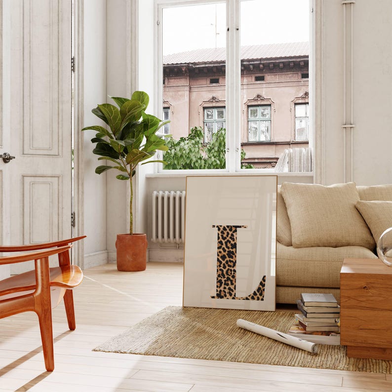 May include: A framed art print featuring the letter "L" with a leopard print design. The artwork is displayed in a bright, airy room with a neutral color palette, a large window, and a potted plant. The room also includes a wooden chair and a beige sofa.