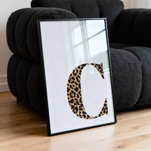 May include: Framed art print featuring a large letter "C" in a leopard print pattern against a white background. The print is encased in a black frame and leans against a dark gray sofa. The artwork adds a touch of wild style.
