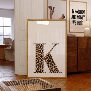 Può includere: Una stampa artistica incorniciata con la lettera "K" con stampa leopardata. La stanza presenta anche altre opere d'arte incorniciate, tra cui una con il testo "IN FASHION AND MONEY WE TRUST". L'opera è esposta in una stanza.
