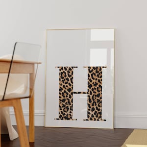 May include: A framed print featuring the letter "H" in a leopard print design against a white background. The gold frame complements the neutral tones of the artwork. The print is displayed on a dark wood floor.