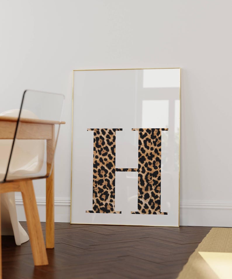 May include: A framed print featuring the letter "H" in a leopard print design against a white background. The gold frame complements the neutral tones of the artwork. The print is displayed on a dark wood floor.