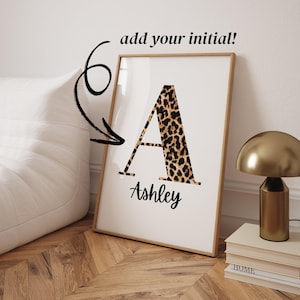 May include: A framed vertical wall art print featuring a large capital letter A with a brown and black leopard print pattern. Below the letter, the name Ashley is written in a black cursive script. The text add your initial! appears above.