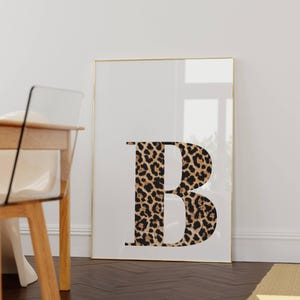 May include: A framed print featuring the letter "B" in a bold, modern font. The letter is filled with a leopard print pattern in shades of brown and black. The frame is gold, and the background is white.