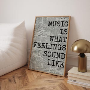 May include: A framed print with the quote "MUSIC IS WHAT FEELINGS SOUND LIKE" in black text. The background is a collage of newspaper clippings. The frame is a light brown color, and the print is leaning against a wall.