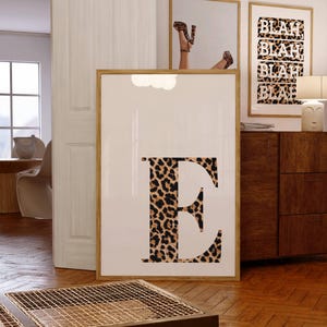 May include: A framed print featuring a large letter "E" in a leopard print pattern. The print is displayed in a room with other framed art, including a mirror reflecting a person wearing leopard print heels. The word "BLAH" is repeated on another print.