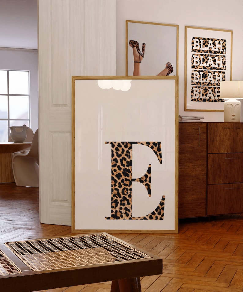 May include: A framed print featuring a large letter "E" in a leopard print pattern. The print is displayed in a room with other framed art, including a mirror reflecting a person wearing leopard print heels. The word "BLAH" is repeated on another print.