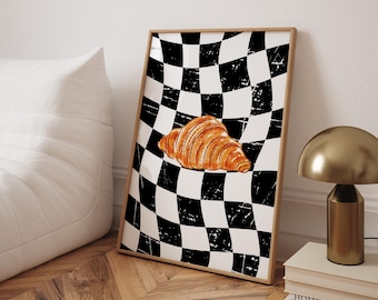 May include: A framed art print featuring a golden-brown croissant on a black and white checkered background. The print has a distressed, vintage aesthetic. The artwork is displayed on a wooden floor, leaning against a white wall.