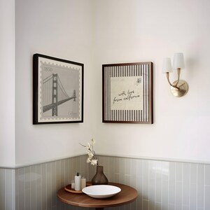 May include: Two square framed wall art pieces featuring a Golden Gate Bridge stamp design and a postcard with the text 'with love from california'. A brass double-arm wall sconce with white shades is mounted next to a round wooden table with a vase.