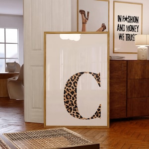 May include: A framed print featuring a large letter "C" in a leopard print design. The print is displayed in a room with other framed art, including one with the text "IN FASHION AND MONEY WE TRUST".