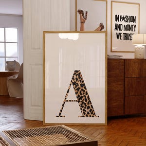 Könnte beinhalten: Ein gerahmter Druck mit dem Buchstaben "A" im Leopardenmuster. Das Bild befindet sich in einem Raum mit anderen gerahmten Kunstwerken, darunter eines mit dem Text "IN FASHION AND MONEY WE TRUST".
