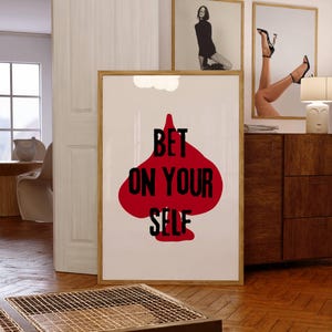 Könnte beinhalten: Ein gerahmter Kunstdruck mit einem roten Pik-Motiv und dem schwarzen Schriftzug "BET ON YOUR SELF". Der Druck wird in einem Raum mit anderen gerahmten Kunstwerken und Holzmöbeln präsentiert.