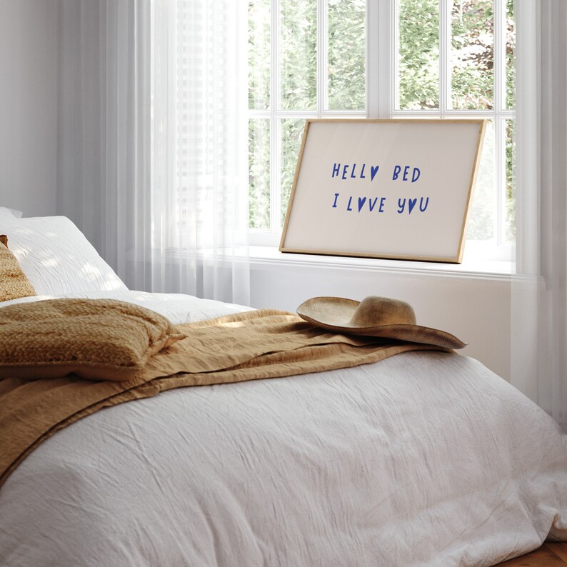 May include: A white bed with a brown blanket and a tan pillow. A straw hat sits on the bed. A framed sign with the text "Hello Bed I Love You" hangs on the wall above the bed.