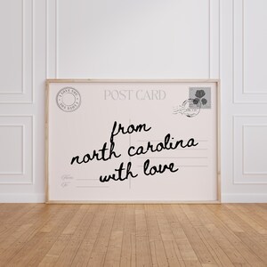 Pode incluir: Impressão emoldurada de um postal com as palavras "from north carolina with love" em escrita preta. O postal tem um fundo rosa claro, um selo postal e um cabeçalho "Post Card". A moldura é de cor de madeira clara.