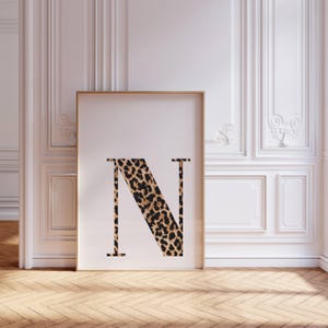 May include: A framed print featuring the letter "N" in a bold, serif font. The letter is filled with a leopard print pattern in shades of brown and black. The frame is a light brown color, and the background is a white wall.