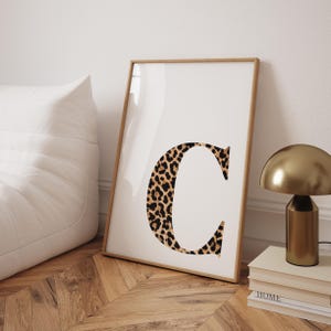 以下が含まれることがあります： ヒョウ柄の「C」の文字が印刷された額装されたプリント。プリントは白い背景にあり、明るい木製のフレームが付いています。