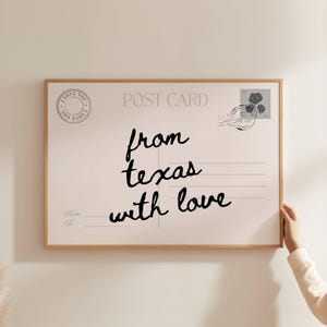 May include: A rectangular wall art print in a light wood frame, designed to look like a postcard. It features the text "POST CARD" at the top, a circular "I LOVE YOU" stamp, a floral postage stamp, and the handwritten text "from texas with love".