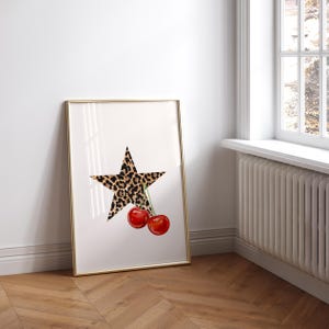 May include: A framed print with a white background featuring a black and brown leopard print star with two red cherries hanging from the star.