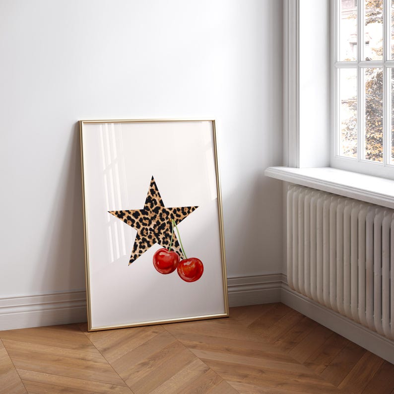 May include: A framed print with a white background featuring a black and brown leopard print star with two red cherries hanging from the star.