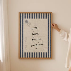 Könnte beinhalten: Ein rechteckiges gerahmtes Wandbild mit einem Vintage-Postkarten-Design auf cremefarbenem Hintergrund und dunkelblauen vertikalen Streifen an den Rändern. Der Text lautet with love from virginia, ergänzt durch eine Briefmarke und einen Stempel.