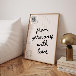 May include: A framed print with the words "from germany with love" in black script. The print has a vintage postcard design with a stamp and postmark. A gold lamp and books are on a wooden surface.