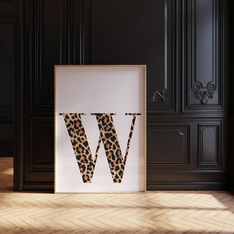 May include: A framed print featuring the letter "W" in a bold, leopard print design. The artwork is set against a white background and framed in a thin, gold-colored border. The print is displayed in a room with dark wood paneling and a wooden floor.