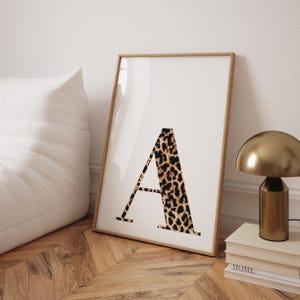 May include: A framed print featuring the letter "A" in a bold, leopard print design. The artwork is displayed in a light brown wooden frame, leaning against a white wall. A gold-colored lamp and books are visible in the background.