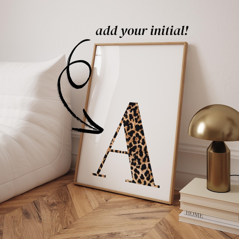 May include: A framed print featuring the letter "A" in a leopard print pattern. The print is displayed on a wooden floor, with a gold lamp and books nearby. Text at the top reads "add your initial!"