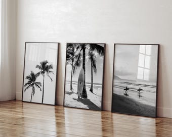 Könnte beinhalten: Drei gerahmte Schwarz-Weiß-Strandkunstdrucke. Die Drucke zeigen Palmen, ein Surfbrett am Strand und Surfer, die am Ufer entlanggehen. Die Rahmen sind dunkelbraun und lehnen an einer weißen Wand.