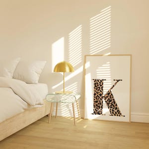 May include: A framed print featuring the letter "K" with a leopard print design. A gold-colored lamp sits on a glass side table with wooden legs. The scene is illuminated by sunlight streaming through blinds.