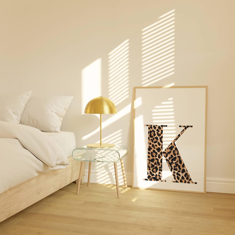 May include: A framed print featuring the letter "K" with a leopard print design. A gold-colored lamp sits on a glass side table with wooden legs. The scene is illuminated by sunlight streaming through blinds.