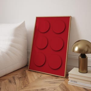 May include: A framed art print with a bold red design featuring six circular shapes. The artwork is in a light wooden frame, leaning against a white wall. A gold-coloured lamp and a stack of books are also visible. The print is a modern design.