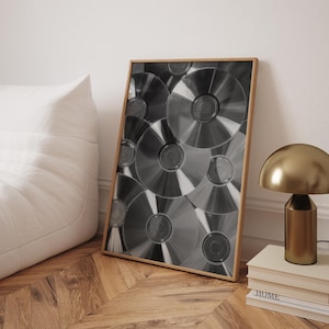 May include: A framed black and white print featuring a close-up of multiple CDs. The CDs are arranged in a layered pattern, creating a textured effect. The frame is a light wood color. A gold lamp and books are on a wooden surface.
