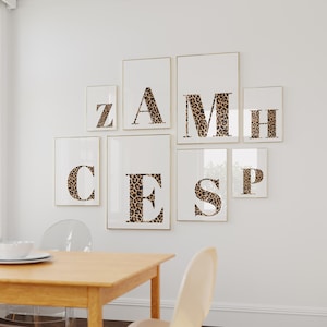 May include: A collection of framed art prints featuring individual letters in a leopard print pattern. The letters are arranged on a white wall, creating a personalized wall decor display. The frames have a gold border.