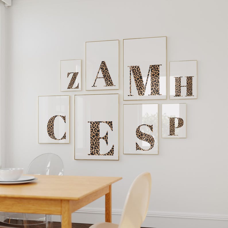 May include: A collection of framed art prints featuring individual letters in a leopard print pattern. The letters are arranged on a white wall, creating a personalized wall decor display. The frames have a gold border.