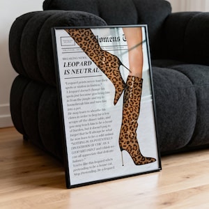 May include: Framed print featuring a fashion illustration of leopard print boots with stiletto heels. The print includes the headline "LEOPARD IS NEUTRAL" and other text. The artwork is displayed against a dark gray sofa.