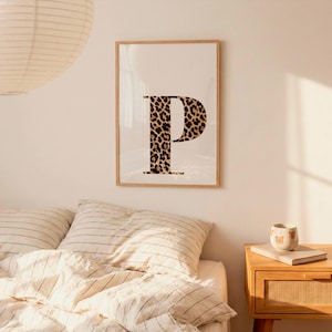 May include: A framed print featuring the letter "P" in a bold, leopard print design. The letter is centered on a cream-colored background, encased in a light wood frame. The artwork adds a touch of wild style to any room.