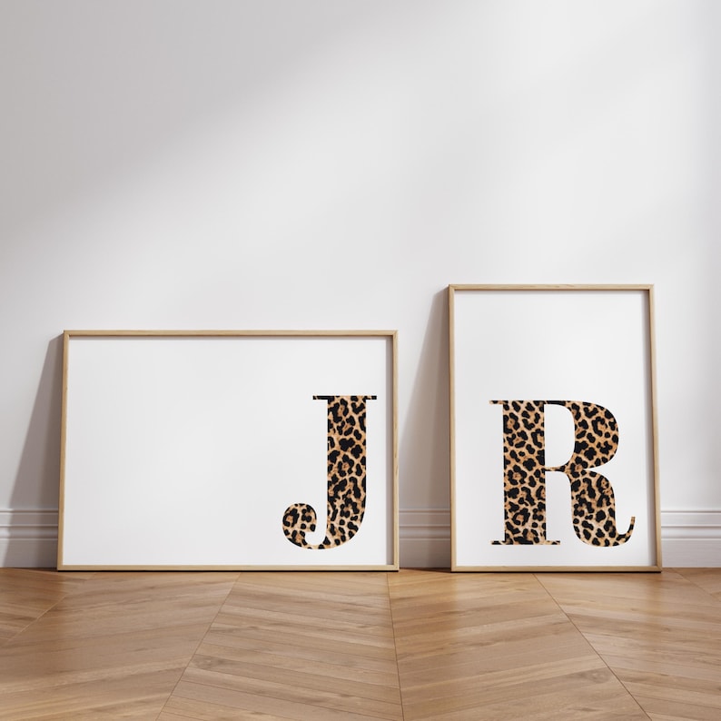 May include: Two framed art prints featuring the letters "J" and "R". The letters are filled with a leopard print pattern, set against a white background. The frames are a light wood color, and the prints are displayed on a wooden floor.