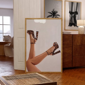 May include: A framed print featuring a pair of legs wearing leopard print high heels. The heels have thick block heels and ankle straps. Two other framed prints are visible in the background, one with a palm tree and the other with legs tied with a bow.