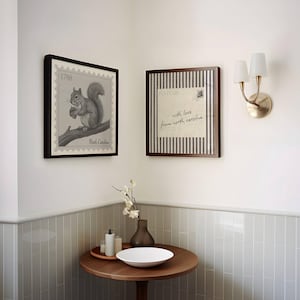 May include: Two square framed wall art pieces featuring a squirrel illustration with the text 1788 North Carolina and a postcard design with the text with love from north carolina, displayed above a round wooden table with a white bowl and vase.