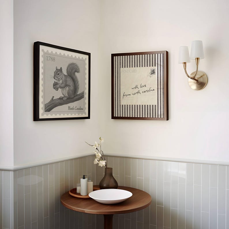 May include: Two square framed wall art pieces featuring a squirrel illustration with the text 1788 North Carolina and a postcard design with the text with love from north carolina, displayed above a round wooden table with a white bowl and vase.