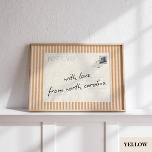 May include: A rectangular framed print featuring a vintage-style postcard design with the text "POST CARD" and "with love from north carolina" in black script. The frame has a light orange and white vertical striped pattern border.