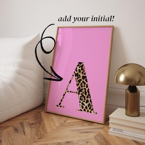 Op de afbeelding: Een ingelijste roze kunstprint met een letter "A" in luipaardprint. De tekst "add your initial!" staat boven het frame. Het kunstwerk wordt gepresenteerd op een houten vloer, met een gouden lamp en boeken op de achtergrond.