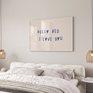 May include: A framed print with the text "Hello Bed I Love You" in blue lettering with heart symbols. The print is hanging on a light beige wall above a white bed with a white and gray patterned blanket.