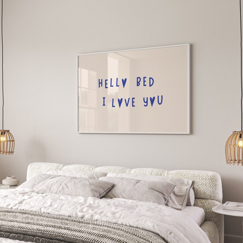 May include: A framed print with the text "Hello Bed I Love You" in blue lettering with heart symbols. The print is hanging on a light beige wall above a white bed with a white and gray patterned blanket.