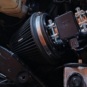 Op de afbeelding: Een motorruimte met een zwart luchtfilter, een zwarte inlaatbuis en diverse metalen componenten. Het luchtfilter is cilindrisch met een zilveren band. Een witte vloeistofreservoir is zichtbaar op de achtergrond.