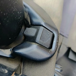 Może przedstawiać: Zbliżenie czarnej plastikowej klamry pasa bezpieczeństwa w samochodzie. Klamra ma teksturowany przycisk zwalniający z pionowymi liniami. Okolice są również czarne i wydają się być częścią mechanizmu pasa bezpieczeństwa.