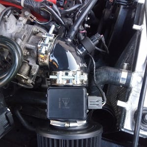 May include: Close-up of a black intake pipe with a silver clamp connecting to a black air filter housing. The air filter housing has a black and silver label that reads "WARRIOR  CARBON 2000".