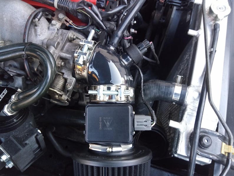 May include: Close-up of a black intake pipe with a silver clamp connecting to a black air filter housing. The air filter housing has a black and silver label that reads "WARRIOR  CARBON 2000".