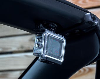 May include: A clear, waterproof action camera case mounted on a black bar. The case is square with a clear front and back, and it has several buttons on the side. The camera is designed to capture video and photos in various environments.
